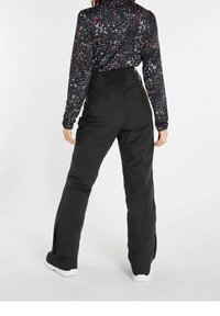 Kobieta w czarnych spodni high-waisted i długim czarnym topie z wielokolorowym wzorem w plamy farby, stojąca tyłem.