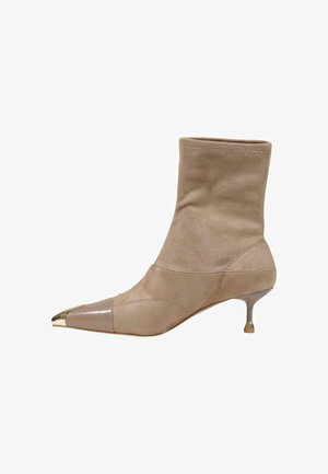 Botín beige de ante con punta afilada que presenta una tapa metálica brillante en beige y oro y un tacón bajo y delgado con punta dorada.