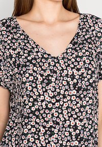 Blouse noire à motifs floraux avec un col en V, des manches courtes, et un petit motif floral composé de fleurs blanches, rouges et bleues sur un fond foncé.