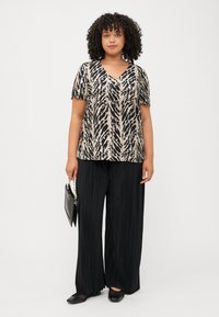 Une femme aux cheveux bouclés se tient confiante, portant un haut à motifs et un pantalon large noir, tenant un sac à main noir.