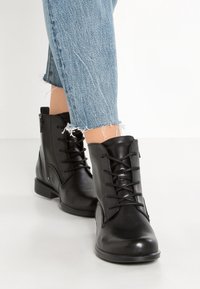 Stivali neri in pelle sopra la caviglia con lacci frontali, punta tondeggiante, tacco basso impilato e texture liscia. Abbinati a polsini in denim rovinato.