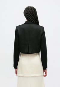 Čierne skrátené sako s hladkou štruktúrou, jedným zadným rozparkom a dlhými rukávmi, doplnené svetlou plisovanou sukňou.