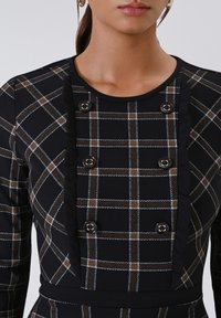 Zwart en bruin geruit jurkje met een ronde hals, ruches aan de voorkant en vier goudkleurige knopen voor een speels detail.