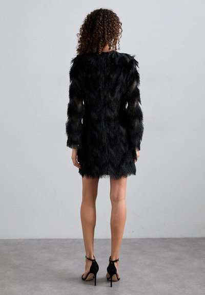 Μαύρο φόρεμα με φτερά και μακριά διαφανή μανίκια με κοφτές λεπτομέρειες. Συνδυασμένο με μαύρες ψηλοτάκουνες γόβες, αποκαλύπτοντας τα πόδια.