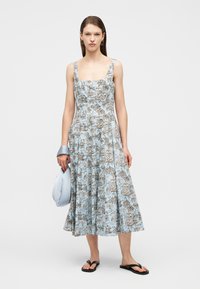 Γυναίκα με αμάνικο μπλε φόρεμα με λουλούδια μήκους μέχρι τη μέση της γάμπας, κρατώντας μια ανοιχτό μπλε τσάντα, φορώντας μαύρα φλατ σανδάλια, στέκεται μπροστά σε απλό φόντο.