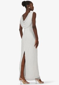 White evening gown with a draped back, embellished with beaded wavy patterns, featuring a thigh-high slit and paired with white strappy heels.