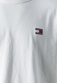 Valkoinen puuvillapaita, jossa on ribbireuna kauluksessa. Paidassa on pieni brodeerattu suorakulmainen logo tummansinisessä, punaisessa ja valkoisessa, jossa lukee "TOMMY JEANS."