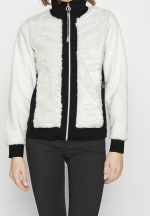 Femme portant une veste polaire à fermeture éclair blanche et noire avec des panneaux avant texturés et des poignets noirs, associée à un pantalon noir.