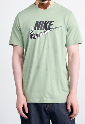 Mann in einem hellgrünen Nike-T-Shirt mit schwarz-weißer, gemusterter Swoosh-Logo, der vor einem schlichten weißen Hintergrund steht.