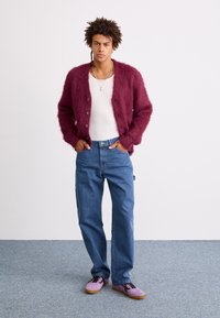 Burgunderfarget, fluffy kardigan over en hvit skjorte, kombinert med blå jeans og lilla sko. Kardiganten har knapper og en avslappet passform.