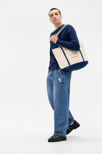 Beige canvas tote bag with navy accents, featuring dual shoulder straps and a printed logo. Worn by a model in a navy garment and jeans.