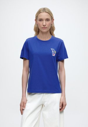 Femme aux longs cheveux blonds portant un t-shirt bleu avec un graphisme de boisson colorée et un pantalon blanc, debout devant un fond uni.