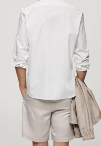 Biała bawełniana koszula z podwiniętymi rękawami, w połączeniu ze jasnobeżowymi szortami. Beżowa kurtka zarzucona na ramię. Prosty, stonowany design.