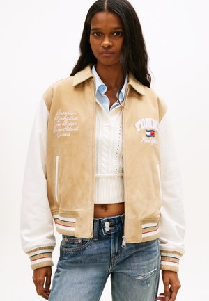 Naine, kes kannab beeži ja valget varsity-jopet ning helesiniseid teksapükse, seisab tavalise valge tausta ees.