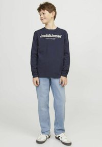 Camiseta de manga larga azul marino con texto de logo blanco, combinada con jeans de color azul claro. El modelo lleva zapatillas negras y blancas, de pie sobre un fondo neutro.