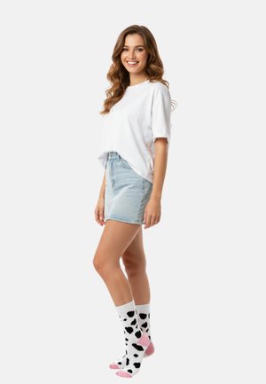Young woman in white t-shirt and light denim skirt smiling, wearing white socks with black spots and pink heels and toes.