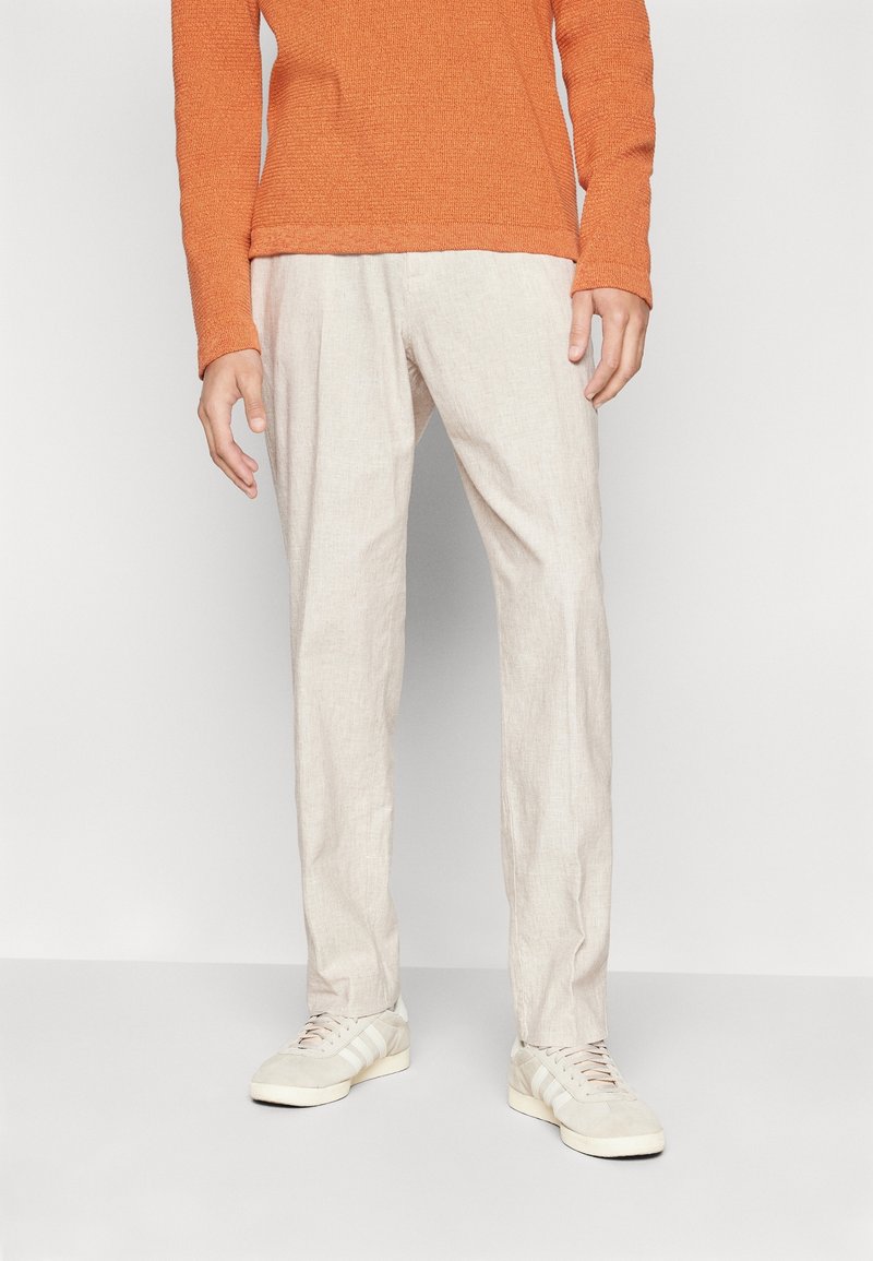 Beige linnedbukser med en afslappet pasform, parret med en struktureret orange sweater og lys grå sneakers med hvide striber.