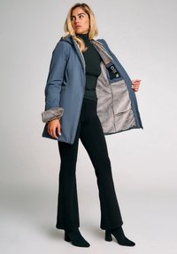 Cappotto grigio imbottito con polsini in pelliccia sintetica, aperto per rivelare un interno morbido di un grigio chiaro. Indossato con un top nero e pantaloni a palazzo, che mettono in mostra un design elegante.