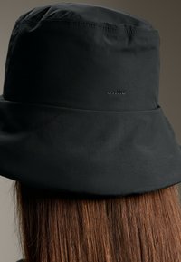 Pălărie neagră tip bucket, confecționată dintr-un material neted, cu un logo subtil pe lateral, având o margine largă și flexibilă. Aspect texturat vizibil.