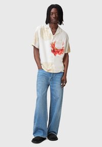 Camisa de manga corta blanca con estampado floral y acentos rojos, con bolsillo en el pecho, combinada con unos jeans de pierna ancha azules y zapatos negros de tipo slip-on.