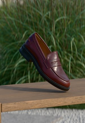 Mocassin en cuir marron foncé avec semelle noire équilibré sur un banc en bois en plein air avec un arrière-plan d'herbe verte haute.