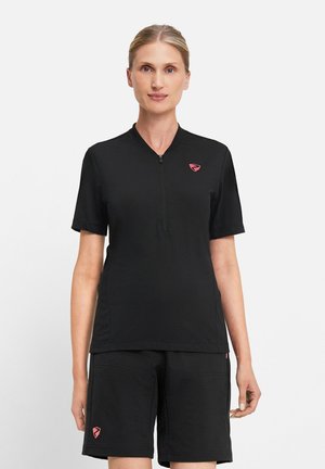 Woman wearing black short-sleeve zippered shirt and black shorts, standing against plain background, facing forward with neutral expression.