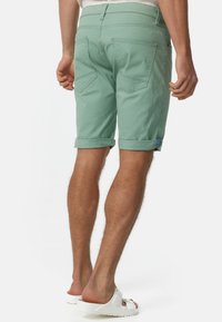 Mann, der helle grüne hochgekrempelte Shorts und weiße Slidesandalen trägt, steht mit dem Rücken zur Kamera vor einem einfarbigen Hintergrund.