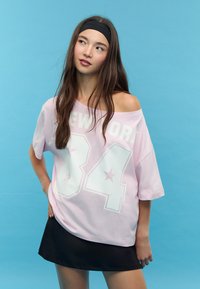Růžové oversized tričko s bílým potiskem "NEW YORK 84" a hvězdnými akcenty, nošené na jednom rameni, v kombinaci s černou sukní.