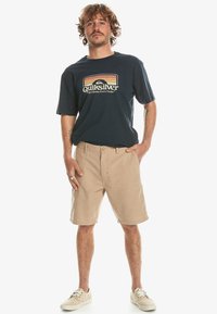 Marinblå t-shirt med retrostil Quiksilver-logga och beiga shorts. Modell som bär ljusa skor, står med händerna på höfterna.