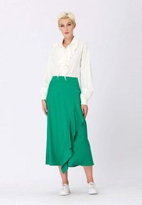 Blouse blanche avec des volants et des manches longues, associée à une jupe plissée verte avec un ourlet à volants. Le modèle porte des baskets blanches.