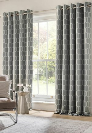 Grey patterned curtains hang on a rod over a large window with a garden view, next to a beige chair and small side table.