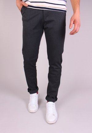 Person iført op rullede mørke slim-fit bukser, en stribet skjorte og hvide sneakers, der går på en ensfarvet baggrund.