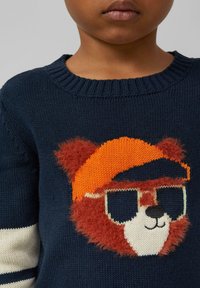 Granatowy sweter z dzianiny z motywem twarzy niedźwiedzia o puszystej, czerwono-brązowej fakturze, pomarańczową czapką i okularami przeciwsłonecznymi. Białe paski na rękawach.