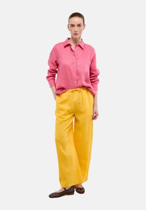Femme portant une chemise rose ample à boutons, un pantalon large jaune et des ballerines tressées marron, debout les mains partiellement dans les poches.