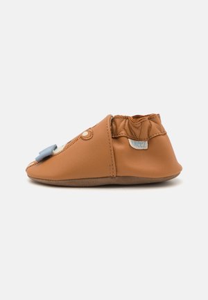 Tan leather toddler shoe with a soft sole, elastic ankle, decorative blue bow, and rounded toe. Features a subtle stitching detail.