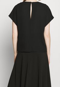 Blouse noire à manches courtes en forme de caps et ouverture en trou de serrure à l'arrière. Confectionnée en tissu lisse avec un design simple et épuré.