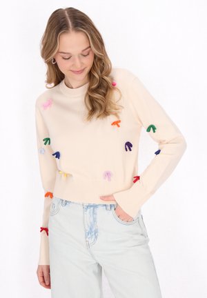 Jasno beżowy sweter dzianinowy z kolorowymi aplikacjami w kształcie kokard z przodu, z prążkowanymi mankietami i dolnym brzegiem, w zestawie z jasnoniebieskimi dżinsami.