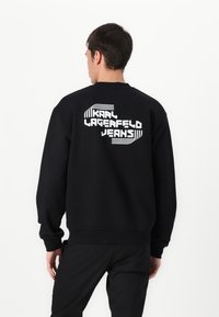 Sweatshirt preto com um grande texto gráfico branco nas costas que diz "KAAI LAGERFELD JEANS", com punhos e bainha canelados.