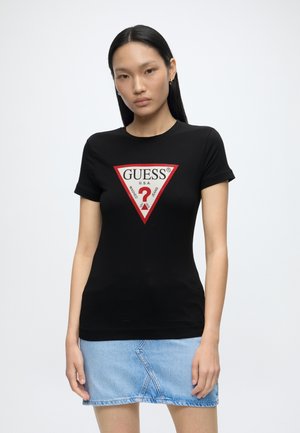 Femme aux longs cheveux foncés portant un t-shirt noir Guess avec un logo triangulaire rouge et blanc et une jupe en jean bleu clair.