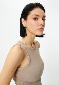 Beige ribbed tank top dengan tali lebar dan leher bulat. Model memiliki rambut hitam pendek dan mengenakan anting-anting bulat. Latar belakang cerah.