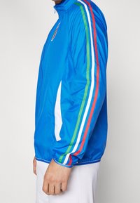 Blå sportjacka med dragkedja fram, prydd med vita ränder och gröna, röda, blå accenter längs ärmarna. Lätt och mjuk textur.