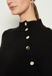 Femme portant un pull noir en tricot avec un col haut fermé par cinq grands boutons dorés sur l'épaule, et des boucles d'oreilles créoles en or.