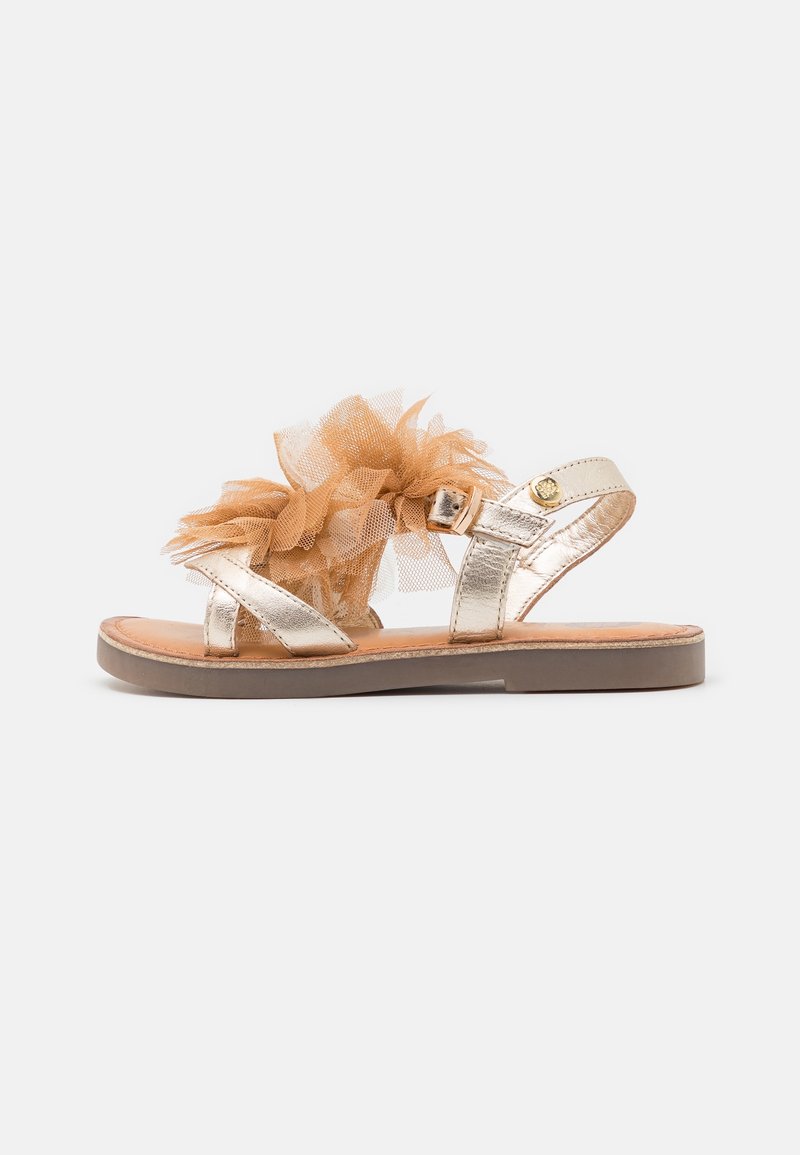 Złote metaliczne płaskie sandały zdobione brązowym tiulowym kwiatem, z paseczkiem na kostkę i klamrą, na beżowej podeszwie, na białym tle.