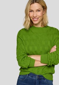 Gestrickter grüner Pullover mit strukturiertem Muster, rundem Ausschnitt und gerippten Bündchen. Kombiniert mit Jeans.