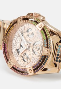 Orologio oro rosa con una lunetta decorata di cristalli luccicanti. Presenta un quadrante trasparente con texture, tre sub-quadranti e un bracciale in metallo con accenti colorati.