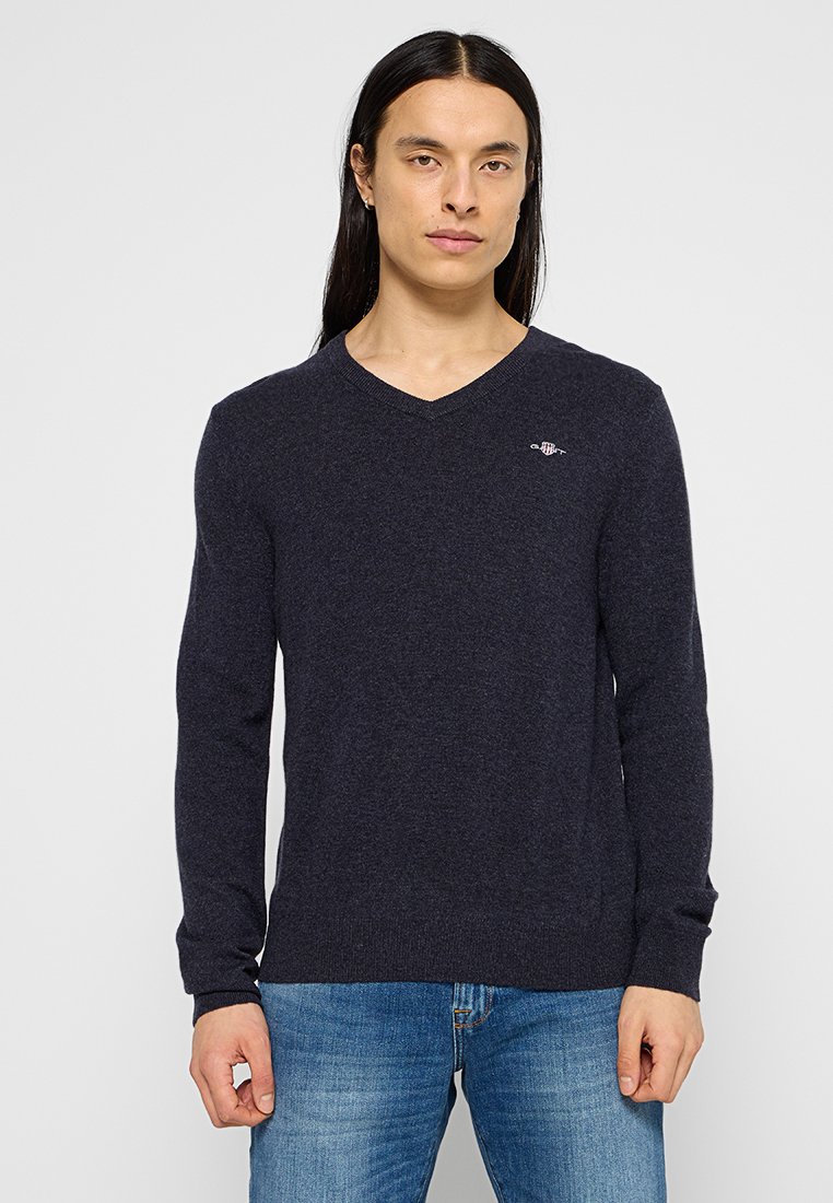 Marineblauer V-Ausschnitt-Pullover aus weichem, strukturiertem Stoff. Mit langen Ärmeln und einem kleinen Logo auf der Brust. Kombiniert mit hellblauen Jeans.