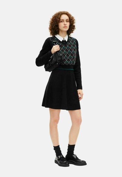 Mulher com cabelo encaracolado, vestindo uma saia preta, colete argyle sobre uma camisa branca com colarinho, segurando uma bolsa preta, de pé com meias e sapatos pretos.