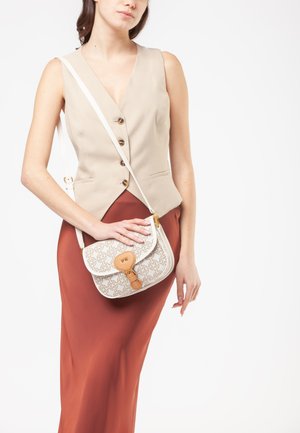 Mujer con chaleco sin mangas beige y falda color óxido, sosteniendo un bolso bandolera blanco con estampado y acentos de cuero marrón claro.