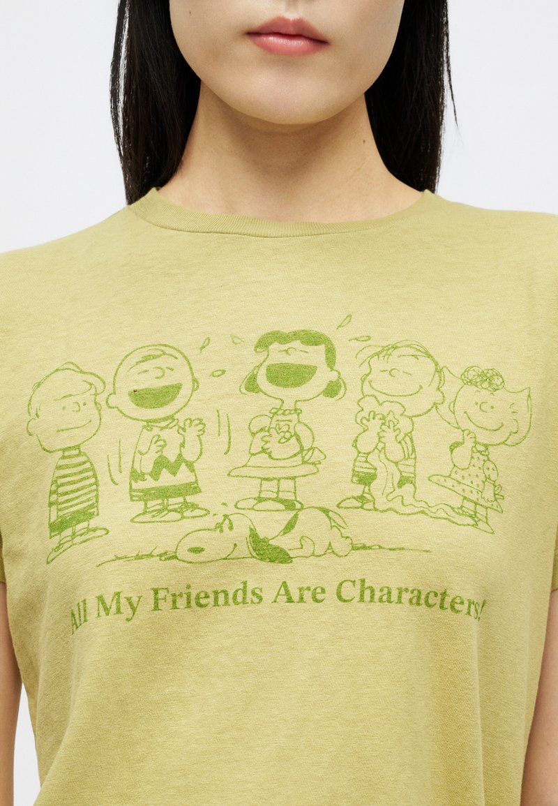 Person som bär en ljusgrön t-shirt med gröna Peanuts-figurer som skrattar och sover, texten säger "Alla mina vänner är karaktärer!"