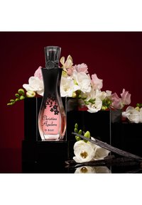 Bottiglia di profumo etichettata "Christina Aguilera By Night" tra fiori bianchi e rosa e bacche di vaniglia su blocchi neri lucidi.